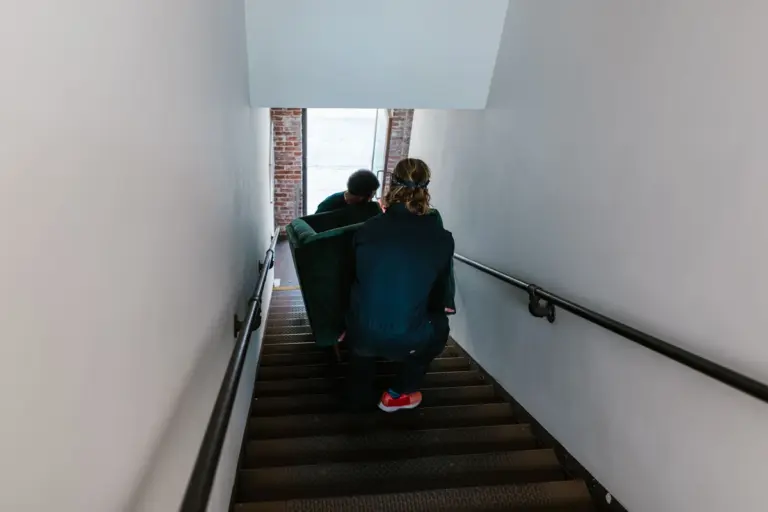 Dos operarios de espaldas bajan un sofá verde por la escalera estrecha de un edificio