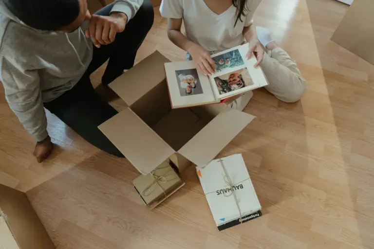 Dos personas revisan un álbum de fotos junto a una caja de cartón abierta y libros atados con cordel